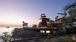 A large house on a hillside overlooking water, with a stone exterior, multiple levels, and a circular structure, featuring outdoor seating areas and a luxurious atmosphere