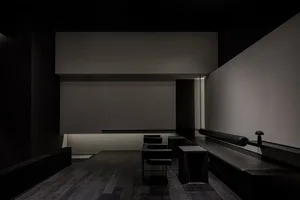 Empty dark room with a black wall and wooden floors, featuring several chairs placed throughout the space