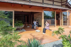 A relaxed social living space with a red house, wooden structure, and people gathered around, some sitting on benches and others standing, with one person using a cell phone