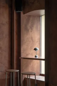 A room with a brown wooden wall, a window, and two lights, featuring a chair near the window, a dining table, and a couch for relaxation