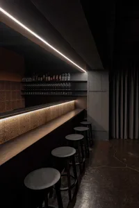 A darkened bar area with a wooden countertop and stools, featuring bottles on display and a cozy atmosphere