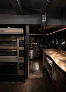 Industrial kitchen with multiple sinks, ovens, and a dining table, surrounded by chairs and kitchen equipment