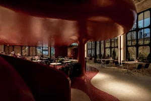 A modern restaurant dining area with multiple tables, chairs, and potted plants, surrounded by red walls and natural light