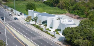 Photograph of a modern building with white walls and a circular shape, surrounded by trees in a natural setting