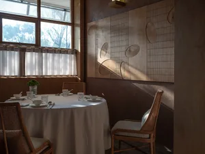 Sophisticated dining area with a white tablecloth, round dining table, chairs, cups, utensils, bowls, and vases