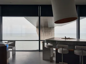 Open modern living space with a kitchen island, stools, chairs, and a large window providing natural light, featuring a marble countertop