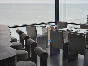 Sophisticated dining area with a large glass window offering a stunning ocean view, surrounded by chairs, a dining table set with wine glasses, cups, cutlery, and bottles