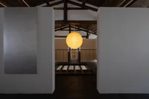 A minimalist room with a white wall and a yellow light hanging from the ceiling, illuminating a staircase leading downwards