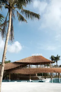Open-air restaurant with a thatched roof, surrounded by palm trees, featuring a large dining area with natural wood and grass elements