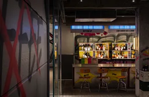A modern bar area with white walls, a metal countertop, and chairs on either side, featuring bottles and a cozy atmosphere