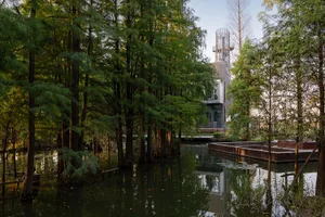 A modern building with a large glass window, surrounded by trees and a serene lakefront setting