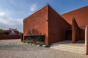 A unique orange building with a red roof and concrete exterior, featuring a large window and two potted plants nearby