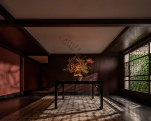 A well-lit interior space with a wooden table, vase of flowers, and potted plants, set against a wooden floor and natural light from a window