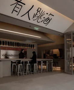 Casual modern restaurant with a bar featuring stools and tables, bottles on display, and a contemporary design