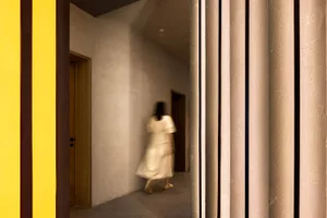 A woman in a white dress walks down a dimly lit hallway with a stone wall and wooden door, flanked by pillars