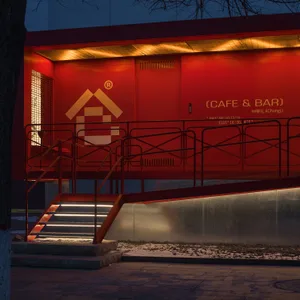A modern bar with a metal staircase leading to a doorway, set against a red building background
