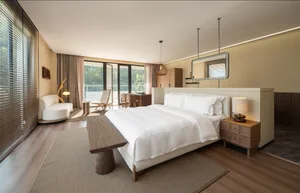 Well-lit bedroom with a large white bed on a hardwood floor, wooden elements, and a dining table in the background