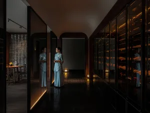 A woman in traditional Chinese clothing stands near a mirror in a dimly lit Asian restaurant with wine glasses and bottles on tables