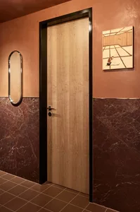A bathroom with a brown marble wall, a wooden door with a metal handle, and a hallway beyond