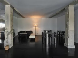 Empty modern living room with black floors and walls, featuring chairs, a dining table, a couch, and a potted plant, with a focus on clean lines and simple design elements