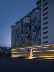 A modern building facade features a large graffiti mural of astronauts in yellow and black tones, set against a concrete or contemporary material background