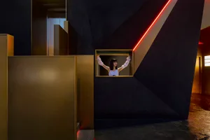 A woman stands in a room with a black wall, looking out the window, surrounded by two other people in a calm atmosphere