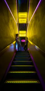 Two women standing on a staircase in a dimly lit room with yellow lights, chairs, and a mix of living and hallway space