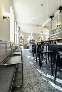 A well-lit restaurant or bar area with a counter and dining table, featuring stools and chairs, and natural light from windows