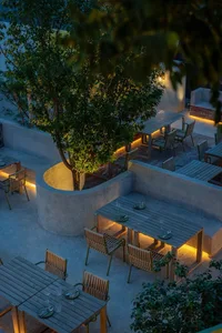 Outdoor dining area with multiple tables, benches, and chairs, illuminated by artificial lights, featuring wood and concrete materials
