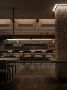 Dimly lit restaurant dining area with a large wooden table and multiple chairs, surrounded by kitchen appliances