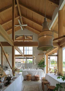 Open living area with wooden tables, chairs, and potted plants, featuring a well-lit and inviting atmosphere with scattered bottles and a comfortable layout