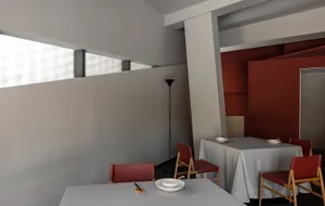 A restaurant dining area with two tables set with red chairs, white tablecloths, and tableware, with a kitchen visible in the background