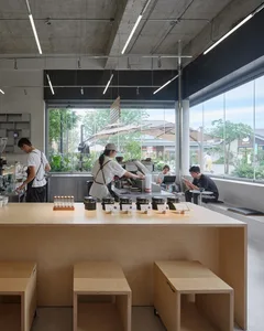 A warm and inviting coffee shop interior with wooden floors and tables, featuring people seated and standing, with cups, bottles, and bowls on the tables