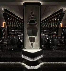 A modern bar area with a triangular structure, black and grey elements, and seating for guests, surrounded by bottles and creating a sophisticated atmosphere