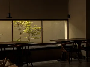 Dining area with two wooden tables and chairs, surrounded by concrete walls and large windows allowing natural light
