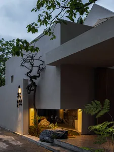 A serene modern building with a large tree in front, its branches extending towards the building, surrounded by people enjoying the outdoor environment