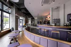 A cozy purple-themed cafe with comfortable seating areas, well-lit by natural and artificial light, featuring unique wall designs and dining tables