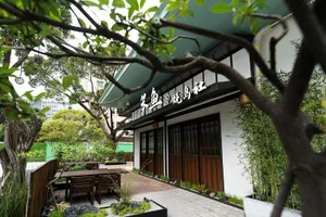 An Asian restaurant with a wooden exterior and green roof, featuring a patio area with chairs and dining tables, surrounded by potted plants