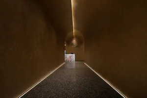 A calm and quiet hallway with a white wall, brown floor, and a large glass box in the center, lit by ceiling lights