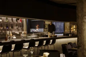 A restaurant bar scene with a counter, wine glasses, and chairs around a dining table, with scattered bottles and an inviting atmosphere