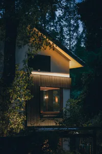 A well-lit modern house at night with a large white building, a wooden door, and two windows, with a chair visible inside