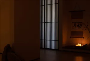 Dimly lit room with a white wall, wooden floor, and Asian artwork, featuring two windows on either side