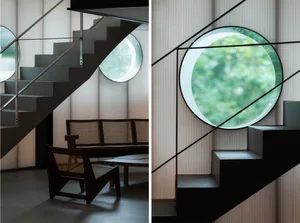 Comfortable living area with chairs and natural light from windows, contrasted by a black staircase railing