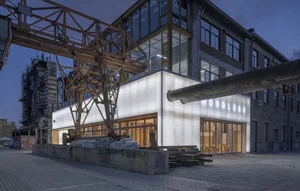 A modern building with a white glass facade and windows, featuring a large crane and chairs in the foreground, set against a night-time urban backdrop