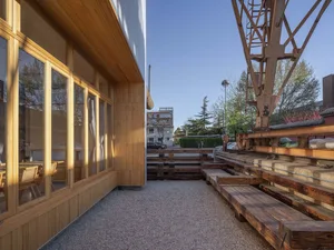 A modern building with wooden walls and a cement ground, featuring large windows and a contemporary design, set against an urban backdrop with parked cars