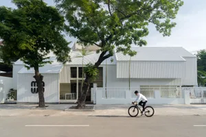 A modern building with a white concrete facade, featuring outdoor seating areas with two chairs and a bench, set in a calm and peaceful environment