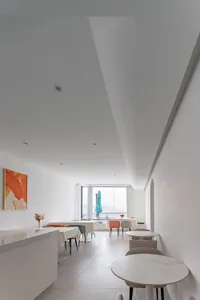 Open living area with a dining table, kitchen sink, refrigerator, couch, chair, potted plant, and vase, surrounded by white walls and natural light