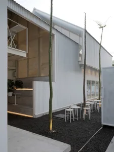 A modern building with a patio area featuring white chairs, dining tables, and potted plants, surrounded by concrete walls and natural light