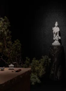 A calm indoor space with a statue on display in front of a black wall, surrounded by potted plants and a cup on a nearby surface