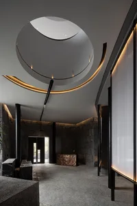 Well-designed hotel lobby with a stone wall, black doorway, and natural light from a circular ceiling window, featuring a bench and potted plants